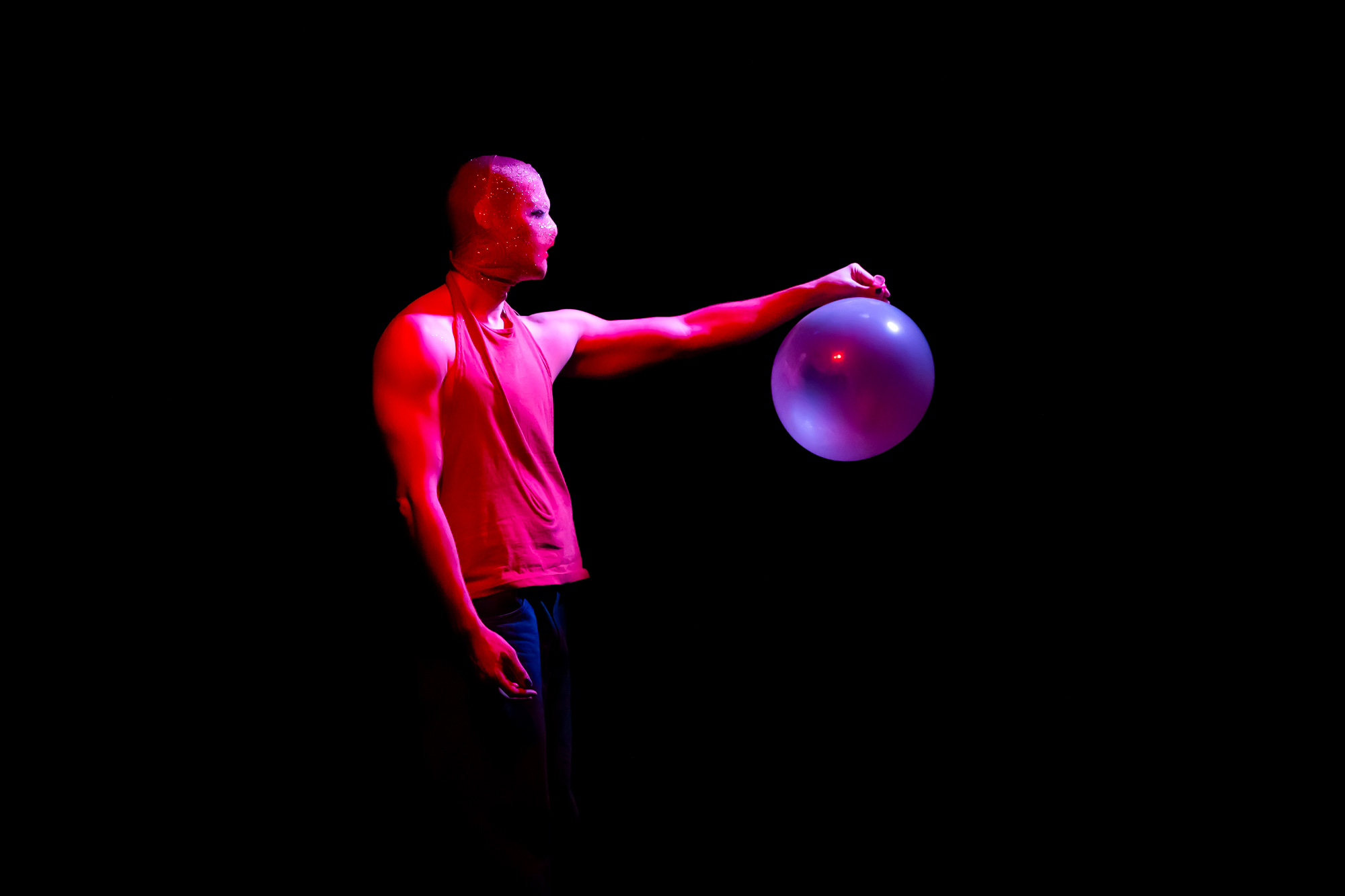 Homme cagoulé tient un ballon de baudruche devant lui, lumière bleue et rose se mélangent.
